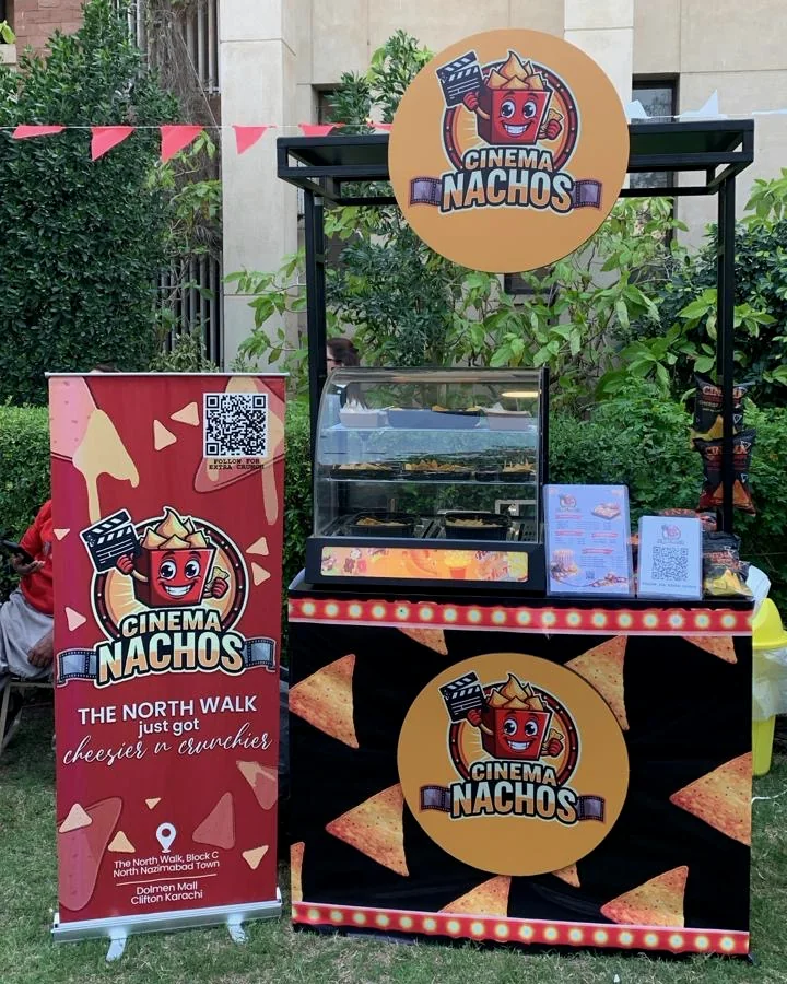 A Cinema Nachos cart set up outdoors at an event: a black counter printed with tortilla chips under the round Cinema Nachos sign, a warm display case of nachos on top, and a Cinema Nachos banner beside it