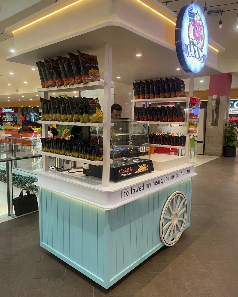 Left-hand view of the Cinema Nachos cart: shelves of Cinema Tortilla Chips under a lit white canopy, the round Cinema Nachos sign and a mint panelled base with a white wagon wheel