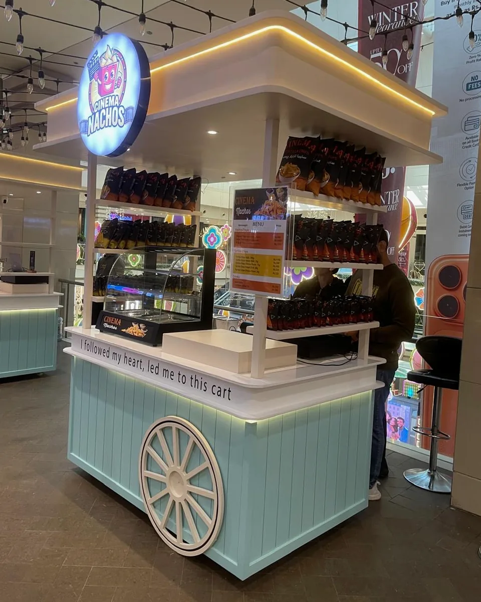 Right-hand view of the Cinema Nachos cart: the menu board and shelves of Cinema Tortilla Chips, string lights overhead, and the mint panelled base with its white wagon wheel